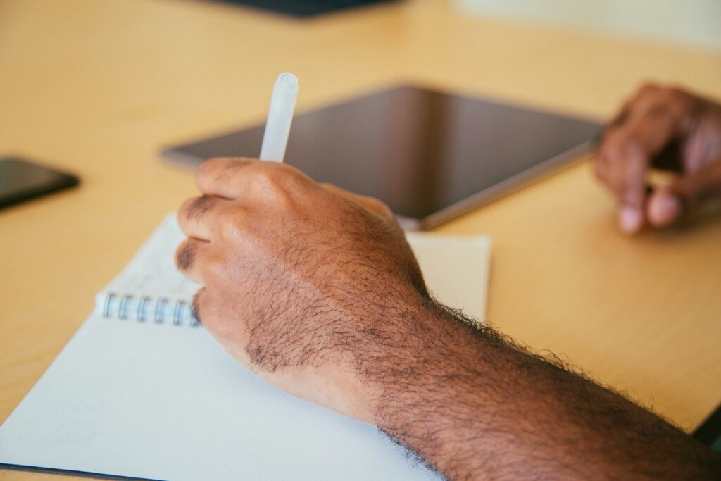 A hand of a person writing in a notebook with a tablet nearby. Provided by Unsplash.